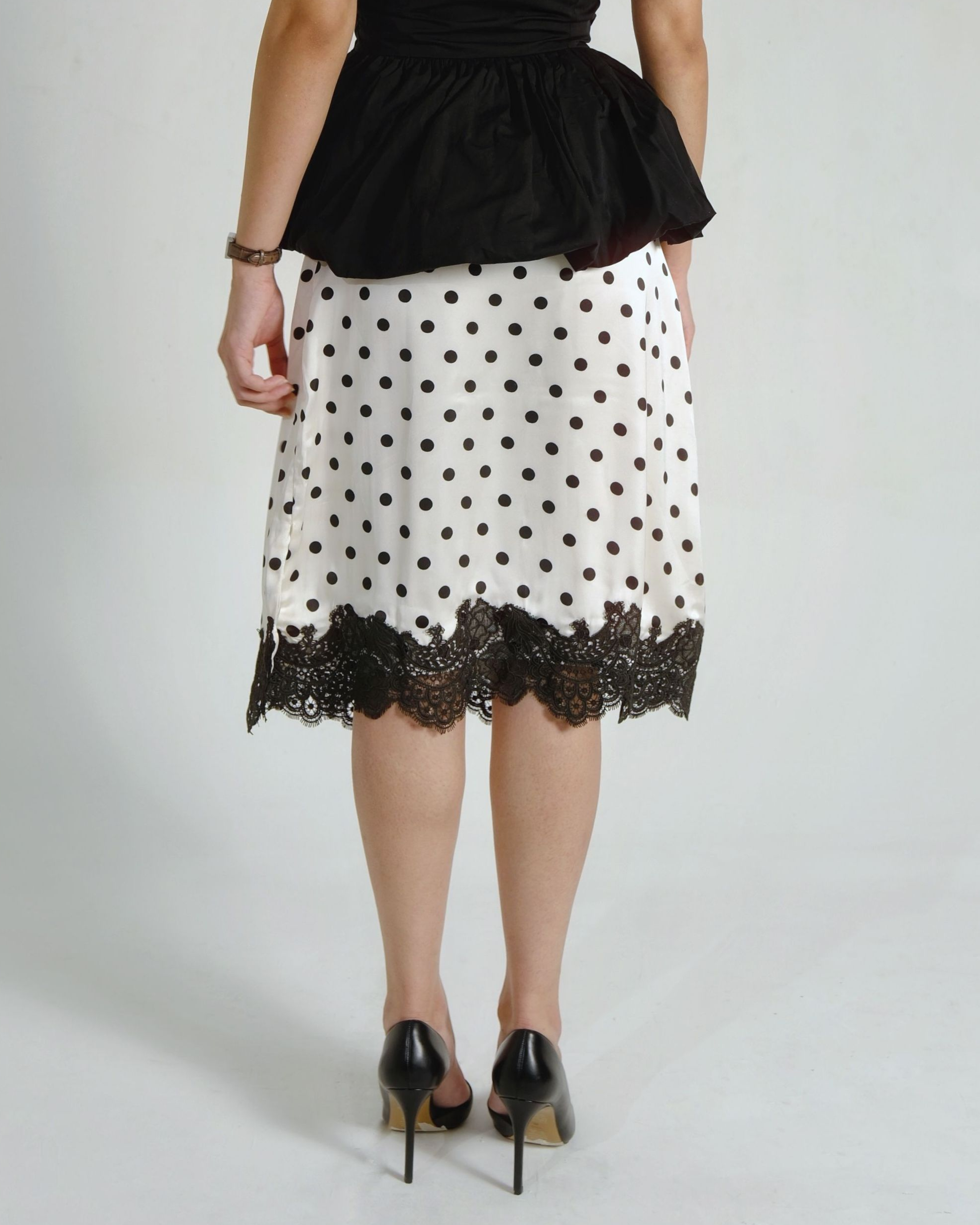 A woman shown from the back wearing a white knee-length skirt with black polka dots and a black lace hem, paired with a black peplum top and black heels, against a light gray studio background.