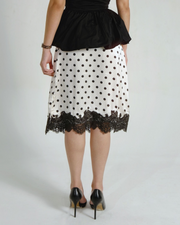 A woman shown from the back wearing a white knee-length skirt with black polka dots and a black lace hem, paired with a black peplum top and black heels, against a light gray studio background.