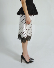 A model is shown from the waist down wearing a white satin midi skirt with black polka dots and a black lace hem, paired with a black peplum top and black pointed-toe heels.