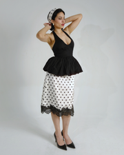 A woman modeling a black halter top with a ruffled peplum hem and a white skirt with black polka dots and a black lace hem, styled with a black and white polka dot headscarf and black pointed-toe heels against a light gray studio background.